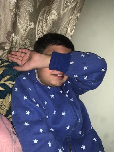 A young child lies on a paisley-patterned cushioned surface, wearing a blue zippered hoodie adorned with white stars. Their face is partially obscured by their left hand, while their right arm extends across the body. Part of their mouth and teeth are visible. A portion of a pink fabric is seen at the bottom right. The background suggests an indoor setting with a light-colored wall.