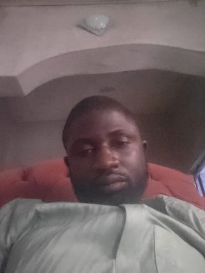 A man with a shaved head and dark skin is positioned close to the camera. He wears a light-colored shirt. The background shows a red cushioned area, possibly part of a sofa or chair. Behind him is a wall with an arched architectural feature and a light fixture. The image is slightly blurry and seems to have been taken in a dimly lit room.