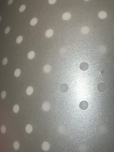 The media file is a close-up, abstract photograph of a textured, metallic surface with a pattern of evenly spaced white dots. The surface appears to be a light gray or silver color, with a slightly rough or matte finish that catches the light in places, creating a subtle shimmer. The white dots are circular and appear to be slightly raised or printed onto the surface, with soft edges that suggest they might be slightly blurred or illuminated. The perspective is angled, giving the impression that the surface is curved or viewed from an oblique angle. There is a bright light source in the upper right quadrant of the image, which causes some of the dots in that area to appear more luminous and even slightly washed out. No people, objects, or discernible scene are visible, only the repetitive geometric pattern. The image is abstract and lacks specific details about time of day, weather, or location, other than the provided context of Letterkenny, Ireland. There is no visible text within the image.