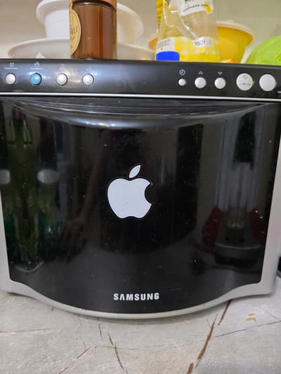 The image shows a close-up of a black Samsung microwave oven with an Apple logo sticker on its top surface. The microwave has several control buttons on its side, some of which are labeled in Cyrillic, including "Разморозка" (Defrost) and "Пароварка" (Steamer). To the left of the microwave, out of focus, are some plastic containers and a glass jar with a label that appears to be in Cyrillic script, reading "любимая крупа" (favorite cereal). The microwave is placed on a concrete surface with some dirt and rust marks, suggesting an indoor setting, possibly a kitchen or utility area. The lighting appears to be artificial, and there are no people visible in the frame. The overall impression is of a used appliance in a somewhat cluttered environment. The Apple logo sticker is a notable detail, indicating a possible modification or personalization of the appliance. The "SAMSUNG" brand name is visible vertically on the right side of the microwave. The text on the label of the jar is partially visible and includes "ikli" and "yor".