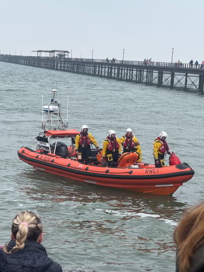 This image captures an RNLI (Royal National Lifeboat Institution) rigid inflatable lifeboat (RIB) with its crew operating on the water beside a long pier, likely Southend Pier, given the provided location context. The scene is observed by people standing on the pier and by spectators in the foreground.

The main subject is the bright orange RNLI lifeboat, prominently featuring "RNLI" branding on its side, along with the identifier "B-826" and the name "JULIE POOLE" near the stern. At least six crew members are visible in the boat, all wearing distinctive yellow waterproof jackets, red life vests with "RNLI" logos, and white helmets. Some crew are seated, while others are standing and appear to be actively engaged in their duties or positioning within the vessel. The boat is equipped with a radar mast and other navigational equipment at its stern, marked "FURUNO" and "NXT".

In the background, the long, dark metal structure of the pier extends across the frame. Numerous individuals are scattered along the pier's length, seemingly watching the lifeboat. Lamp posts are also visible on the pier. The water is a choppy grey-green, suggesting a slightly overcast day with no bright sunshine.

In the immediate foreground, parts of two spectators are visible, adding to the sense of an ongoing event or public viewing. One person with blonde braided hair wearing a dark jacket is seen in the lower right, and another person's head is partially visible in the upper right.

The overall activity suggests the RNLI lifeboat is either conducting a training exercise, performing a patrol, or transiting near the pier, attracting public attention.