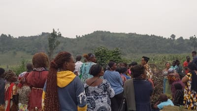 A group of people, mainly women, stand outdoors against a background of rolling hills. The women are dressed in a variety of casual clothing, some with colorful patterns. Most of the group is looking towards a central point within the gathering. The landscape includes a field of crops, possibly corn, suggesting an agricultural setting. The sky is overcast, casting a soft light over the scene.