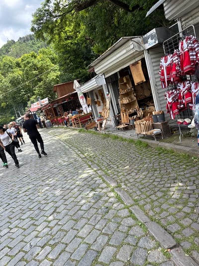 The image shows a cobbled street in Bachkovo, Bulgaria, lined with small shops selling souvenirs. The shops are open, displaying a variety of items including traditional Bulgarian clothing with red and white embroidery, wooden crafts, and other trinkets. In the foreground, several people are walking along the street. One man in a black t-shirt and black pants is walking away from the camera, with two women in white tops and black pants walking in front of him. The street is paved with cobblestones and has grass growing between them. In the background, there are trees and a hillside covered in greenery. The shops have awnings and some have tables and chairs set up outside, suggesting they might also be cafes or restaurants. The overall atmosphere appears to be that of a busy tourist area.