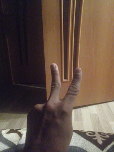 The image is a close-up, slightly blurry, low-light photograph of a person's hand holding up two fingers in a peace sign. The hand is in the foreground, partially obscuring the background. The background appears to be an interior scene with a wooden door or cabinet with vertical paneling. There is also a glimpse of a floor with a patterned rug. The lighting is dim and warm, casting shadows. The scene is not clearly identifiable as being in Shakhtinsk, Kazakhstan.