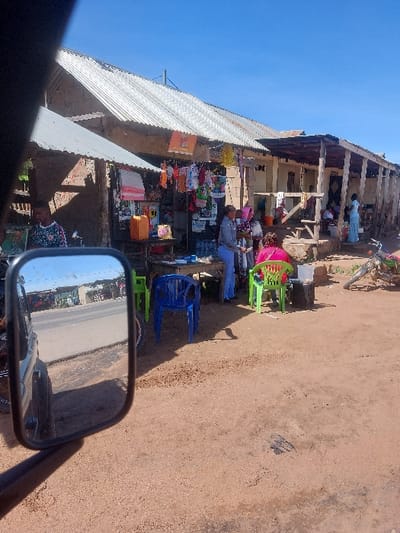 The scene depicts a street market in Kimamba, Tanzania, captured from the interior of a vehicle with a prominent side mirror in the lower-left foreground. The ground is an unpaved reddish-brown dirt surface, showing tire tracks and small divots.
In the mid-ground, a row of single-story commercial buildings with corrugated metal roofs runs horizontally. The buildings feature light-colored, possibly stucco or mud-brick, walls and extended wooden-post verandas or awnings. Several market stalls are set up in front of these structures.
On the left, a stall displays various items, including hanging clothes (infantwear, t-shirts) and what appears to be textile goods. An orange jerrycan and other containers are visible on a nearby wooden table. A male individual in a dark patterned shirt is partially visible on the far left, standing near a market display.
Towards the center-right, a woman in a light blue long-sleeved top and dark pants stands near a table with items. Another woman, wearing a pink top, is seated on a bright green plastic chair, facing away from the camera. A blue plastic chair stands empty nearby. Other individuals are visible further back under the building's overhangs, including a woman in a light blue dress on the far right. A bicycle is leaned against a structure on the right side.
The sky is clear and bright blue. The vehicle's side mirror reflects a paved road, distant buildings, and what appears to be other vehicles or motorcycles. Sunlight casts clear shadows, indicating midday conditions.