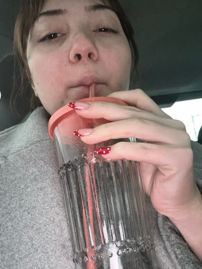 A young woman with light skin drinks from a clear, ribbed cup with a light pink lid and straw. Her hand, adorned with red and white polka-dot French-tipped nails, grips the cup. She wears a gray sweater, and a car window is visible in the background. Her face is turned toward the camera as she sips her drink.