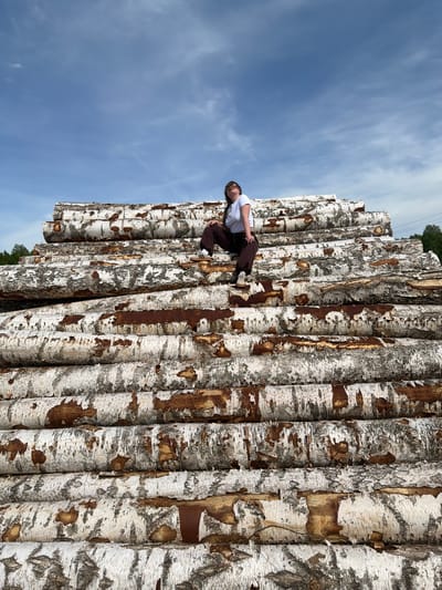 A young woman is sitting on a stack of birch logs. The logs have white bark with brown markings and are stacked horizontally. The woman is wearing a white t-shirt and brown pants. The sky is blue with white clouds and visible in the upper left of the image. Green trees are visible in the top center of the image. There are power lines in the upper left of the image. Based on the presence of birch logs and trees, this image appears to be taken in a rural or wooded area. Based on information provided, the location is Ural'skoe, Russia.