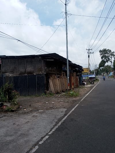 This image depicts a roadside scene in Khajuripada, India, under an overcast sky. In the foreground, a dark asphalt road with a solid white shoulder line runs diagonally from the bottom right towards the upper left. To the left of the road is an unpaved, uneven dirt and gravel shoulder with sparse green vegetation, including potted plants and weeds.
Adjacent to the shoulder, on the left, is a single-story building constructed with dark corrugated metal sheets forming the lower exterior walls and lighter, possibly woven bamboo or straw panels for the upper sections. The building has a dark corrugated metal roof. Stacked vertically against the front of this building are numerous light-colored wooden frames or panels, suggesting a carpentry or building material shop.
A tall, grey metal utility pole stands directly on the shoulder near the center of the image, with multiple overhead electrical wires extending across the frame. Further down the road, another utility pole, dark brown and appearing to be made of wood, is visible. Beyond the immediate building, other small structures and signs can be seen along the roadside, including a blue and yellow sign in the midground.
On the asphalt road, a lone individual on a blue scooter is traveling away from the viewer towards the upper right. Further in the distance, other vehicles are barely discernible. A scooter is parked on the dirt shoulder near the blue and yellow sign. Large, leafy green trees are present in the background, particularly on the right side of the road. The overall environment suggests a semi-urban or rural commercial strip.