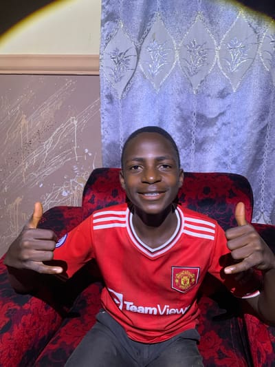 The media file is a portrait of a young Black male, likely a teenager, wearing a red Manchester United football jersey. He is seated on a red couch with a dark, floral pattern. The background features a light-colored curtain with a diamond-shaped pattern in a lighter shade and what appears to be a painted wall with white and brown splatters. The lighting suggests it is nighttime, with a strong light source creating a halo effect on the left side of the image. The individual is smiling and giving a thumbs-up gesture with both hands, conveying a positive and enthusiastic emotion. The visible text on the jersey includes the Manchester United crest and the words "TeamView". The image is oriented vertically, as if taken with a phone in portrait mode. The setting appears to be indoors, possibly a living room, in Barakin Sabawa, Nigeria.
