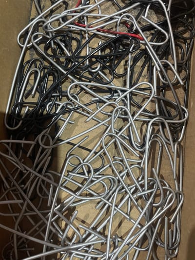 The image is a close-up, overhead shot of a disorganized pile of metal paper clips. The paper clips are primarily silver, with a significant number of black paper clips mixed in. There is also a small section of a red paper clip visible. The paper clips are in a state of disarray, overlapping and tangled, filling the frame. The background appears to be a cardboard box, indicated by the textured brown surface. The lighting is somewhat dim, casting shadows that highlight the metallic sheen of the paper clips. There are no people or any other objects present. The scene is devoid of any context related to the city of Сокуры, Russia, or any specific location within it.