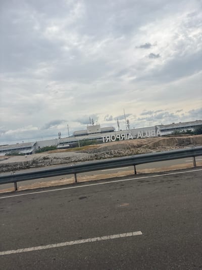 The image captures a wide view of an airport terminal building with prominent signage. The perspective is from a road adjacent to the airport, with the road occupying the right side of the frame. The sky is overcast, suggesting a cloudy day, possibly in the morning or late afternoon given the lighting.
The airport building itself is modern, with a long, multi-story structure visible. Antennas and communication towers are mounted on top of the building, indicating its functional purpose. The "ABUJA AIRPORT" sign is clearly displayed in large, white, capitalized letters, positioned vertically along the side of the building and clearly visible. The surrounding landscape appears somewhat arid or dry, with patches of vegetation and dirt.
The road is paved asphalt, marked with white lines. A metal guardrail separates the road from the airport grounds. The overall scene is one of an active transportation hub under a cloudy sky. There are no people or vehicles prominently visible, focusing the viewer's attention on the architecture and signage of the airport. The time of day is not definitively clear, but the diffuse light from the clouds suggests it is not midday with direct sunlight. The location is explicitly identified by the signage as Abuja Airport, Nigeria.