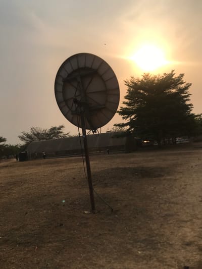 The image, captured in Umudike, Nigeria, depicts an outdoor scene bathed in the dramatic light of a low sun, suggesting either sunrise or sunset. A large, circular satellite dish, light grey in color, stands prominently on a brown pole in the left foreground.
The sun, a brilliant white-yellow source of intense light with visible rays and significant glare, dominates the upper left portion of the sky. This powerful light creates a hazy, warm, and somewhat diffused atmosphere, casting a warm tone across the entire scene.
To the right of the satellite dish and extending into the mid-ground are several dark green, tall, and slender trees, resembling conifers, their forms partially silhouetted against the bright sky. The ground, occupying the lower and right sections of the frame, is a vast expanse of dry, textured, light brown earth, indicative of a dusty or arid environment.
In the distant background, behind the trees, a long, low-lying structure, possibly a greenhouse or an agricultural building, stretches horizontally across the scene. Small, indistinct figures are faintly visible near this background structure. No specific activity is clearly discernable, and there is no visible text. The overall impression is an expansive, rural or semi-agricultural landscape under a powerful sun, with a slightly hazy atmospheric condition.