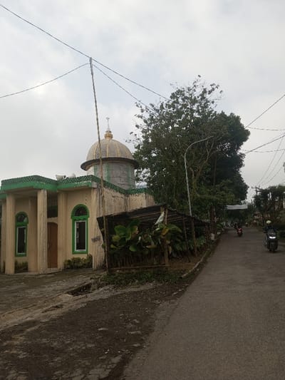 A small church-like structure with a dome stands near a road. The building is beige with green trim. A makeshift wooden lean-to structure sits between the building and the road, with foliage underneath. The road curves into the distance where several people on motorcycles are seen. Power lines cross the sky above a few trees, adding to the urban landscape. The sky is overcast.