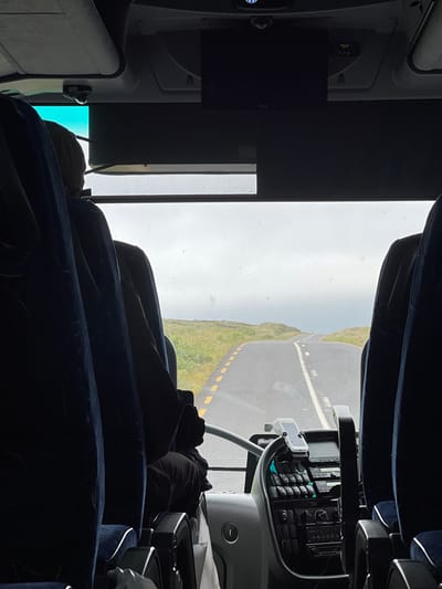 The image shows the view from inside a bus, looking forward through the windshield. The seats of the bus are in the foreground, with a person seated in the driver's position, partially obscured by the seats and dashboard. The road ahead is paved and has yellow dashed lines marking the lanes. The road winds through a grassy, hilly landscape under an overcast sky. The scene is consistent with the rural landscape of Meenacreevagh, Ireland.