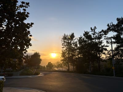 The image shows a street in Aliso Viejo, California, USA, during sunset. The sun is setting behind rolling hills in the distance, casting a warm glow over the landscape. Silhouetted trees line the street, and a speed limit sign reads "SPEED LIMIT 25". In the foreground, a paved road curves to the right, with a sidewalk and manicured landscaping on the left. The sky is a gradient of blue and orange, with a few wispy clouds. There are no people or animals visible in the image.