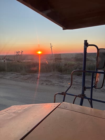 The image captures a sunset scene from the perspective of what appears to be a vehicle or piece of heavy machinery. The sun is a bright orange orb low on the horizon, casting a warm glow and creating a lens flare that extends upwards. The sky transitions from a pale blue at the top to vibrant orange and pink hues near the horizon. In the middle ground, there is a grassy, uneven terrain with some wooden planks scattered on the ground. Two wooden towers with electrical equipment are visible on the left, and utility poles with wires stretch across the landscape. The foreground is dominated by the metal surface of the machinery, which is reflecting the sunset's colors. A blue metal frame with mirrors is also visible, suggesting the vantage point is from a driver's cab or an operator's station. The overall environment appears to be an open, possibly industrial or rural area on the outskirts of Tatsinskaya, Russia, characterized by dusty ground and sparse vegetation.