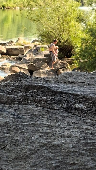 A man, shirtless, with shorts, is holding a baby in a carrier against his chest. They are standing on rocks near a body of water. The water is green and reflects the surrounding trees. There are more rocks and vegetation in the background. The foreground is dominated by large, rough rocks. The image appears to be taken in an outdoor setting, possibly a river or lake in Enns, Austria, given the context provided.