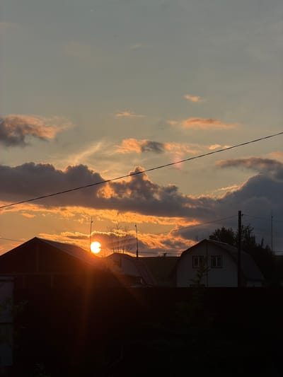 This is a vertically oriented, landscape-oriented photograph taken at sunset in Emel'yanovo, Russia. The scene depicts a rural or suburban setting with a focus on the dramatic sky.
The sky is filled with a mix of dark, brooding clouds and wispy, illuminated clouds tinged with orange and pink hues, indicative of the setting sun. A single, bright streetlamp is visible, its light a prominent focal point in the middle right of the frame, casting a warm glow. The sun itself is a bright, intense orb peeking through the clouds, its rays emanating outwards.
In the background, two houses are partially visible, silhouetted against the colorful sky. The house on the left appears to be a lighter color with visible windows, while the structure on the right is almost entirely in shadow. Power lines are strung across the upper portion of the image, adding to the sense of a lived-in, developed landscape. The overall mood is serene and somewhat melancholic, characterized by the fading light and the quiet presence of the houses. The time of day is clearly sunset, and the weather appears to be clear with dramatic cloud formations. No visible text is present in the image.