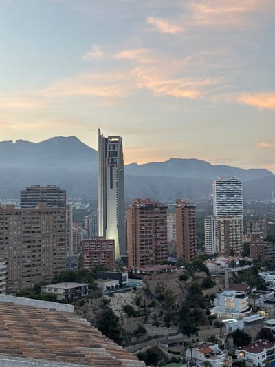 This is a vertically oriented, eye-level, long shot image of a coastal city with a mountainous backdrop, likely taken at dusk. The sky is a soft gradient of pale blue and warm, peachy hues, with wispy clouds suggesting the transition from day to evening.

The foreground is dominated by a textured, reddish-brown tiled roof, angled from the bottom right corner upwards. Beyond this roof, the scene opens up to reveal a densely built-up urban landscape cascading down a steep slope towards the sea. Numerous buildings of varying heights and architectural styles are visible, including modern high-rises with reflective facades, and older, more traditional apartment blocks with balconies. Some of the buildings are illuminated, indicating it is nearing nighttime.

The city is nestled against a rugged, grey mountain range that stretches across the middle ground and forms the distant background. The slopes of the mountains are partially covered in sparse vegetation. The overall impression is of a bustling Mediterranean city built on a dramatic incline, bathed in the soft light of sunset. The scene is devoid of people, with the focus entirely on the urban and natural environment. No text is visible in the image.