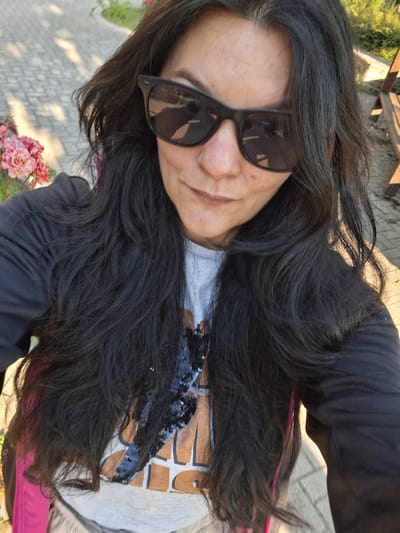 A medium shot selfie features a woman with long, dark, wavy hair and dark sunglasses. She is wearing a black jacket over a light-colored t-shirt with a graphic print and some sequined embellishments. Her hair is partially obscuring her face. In the background, a paved walkway, a wooden bench, and pink flowers are visible. The scene suggests an outdoor setting, possibly a park or garden. The sunlight indicates it is daytime. The background appears to be a paved area with cobblestone or similar material, common in older European city centers. The presence of a bench suggests a public space. The overall lighting indicates a sunny day. The woman is facing the camera, taking a selfie. The focus is on the woman, with the background slightly blurred.