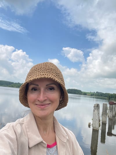 A woman is taking a selfie outdoors with a body of water and a cloudy sky in the background. She is wearing a light-colored hat and a tan jacket over a pink and gray top. The water appears to be a lake or a wide river, with several old concrete pillars visible in the water to her right. The background shows a treeline across the water. The image is a close-up of the woman, with the background out of focus. The location is identified as Bereznik, Russia.
