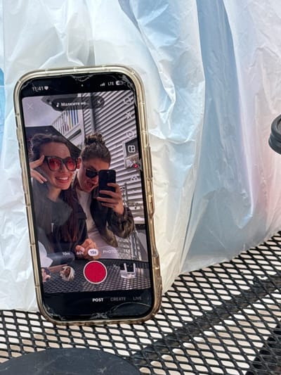 A smartphone with a clear case and a visibly cracked screen is resting on a crinkled, light-colored surface, possibly a white plastic bag. To the right, a black mesh pattern suggests a table or chair. The phone's screen displays a selfie-style view of two young women, both smiling broadly. One woman, wearing sunglasses and with her hair pulled back, is taking the picture or video, holding up another smartphone. The second woman, also in sunglasses, smiles at the camera. The background of their selfie appears to be an indoor setting with parallel lines, possibly an escalator or building interior.
The phone's interface indicates it's likely a social media or video recording application, showing options like "15s," "60s," and "10m" for video duration, as well as "PHOTO," "TEXT," "POST," "CREATE," and "LIVE" functions. A large red circular record button is prominently displayed. Visible text on the screen also includes the time "11:41," "LTE" for cellular service, and "44" (likely battery percentage). On the left side of the screen, there's text in Cyrillic, "Малките не..." (meaning "The little ones are not..." in Bulgarian), next to a music note icon, suggesting a music selection or caption. A heart icon is visible in the bottom left. The women appear happy and engaged in recording their interaction. The image provides no specific location cues for Hampton, United States of America, beyond the device itself.