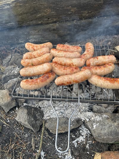A close-up, top-down view captures sausages grilling over an open fire. The scene appears to be outdoors, possibly in a rustic or camping setting, with a log overhead and rocks surrounding the fire pit. The sausages, varying in shade from light pink to golden brown, are arranged on a metal grill rack. Wisps of smoke rise from the grill, indicating the cooking process. The ground is covered in ash, small stones, and pine needles, suggesting a natural environment. The image does not depict any people. The location in Chmyrivka, Ukraine, is not visually evident from the image itself.