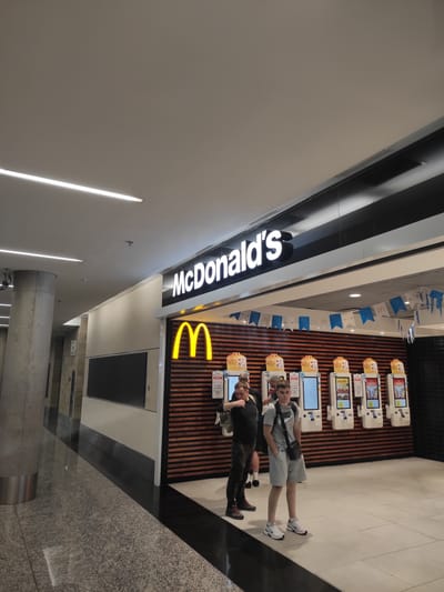 In this image, two young men are seen standing in front of a McDonald's restaurant. The McDonald's logo, a large yellow "M", is prominently displayed on the right side of the frame, with the "McDonald's" text in white illuminated lettering on the left. The restaurant interior features numerous self-ordering kiosks, some of which are displaying menu items. The overall setting appears to be a modern, well-lit space, likely within a public area given the presence of what appears to be airport signage and flooring. The men are casually dressed, with one wearing a grey t-shirt and shorts, and the other in a black t-shirt and pants. They seem to be interacting with the kiosks or waiting in line, though their expressions are neutral. The image is taken from a slightly elevated angle, looking down towards the subjects and the restaurant. The background shows a wall with vertical wooden slats, characteristic of some McDonald's branches. Decorative flags with the McDonald's logo are hung along the top of the wall. The lighting suggests it could be daytime, but the indoor setting makes it difficult to determine the exact time of day or weather conditions. The image provides a glimpse of a typical fast-food experience in a busy environment.