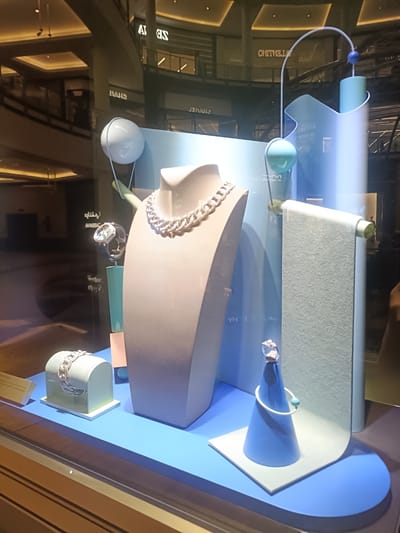 A brightly lit jewelry store window display is presented, exhibiting several pieces of high-end jewelry against a backdrop of minimalist, pastel-toned sculptural elements. The scene is within a modern shopping mall, identifiable through reflections on the glass.
The display rests on a rectangular, bright blue platform. From left to right:
1. A white bracelet with an intricate, possibly diamond-studded, woven design is presented on a light blue arched stand with a white base.
2. Behind and to the left of the bracelet, a wristwatch with an ornate dial and a light-colored strap is displayed on a small, light blue rectangular block.
3. Centrally, a substantial, multi-strand white metal necklace, heavily encrusted with small, sparkling stones (likely diamonds), is showcased on a beige mannequin bust.
4. To the right, a ring featuring a prominent central gemstone is positioned atop a light blue conical stand. A thin white wire-like bracelet with a small turquoise spherical bead is draped around this cone.
Decorative elements in the display include various geometric shapes and spheres in shades of pastel blue, light green, and white, along with small pink and light blue rectangular blocks. Thin white curved rods connect some of these elements, creating an abstract, artistic arrangement. A tall, textured light gray-blue panel hangs vertically on the right side of the display.
The reflective surface of the display window reveals the inverted interior of a high-end, multi-story shopping mall. Clearly visible reflections include the inverted names of luxury brands, specifically "VALENTINO" and "CHANEL," strongly indicating the location is in a premium retail environment consistent with large malls in Dubai, United Arab Emirates. The mall's interior shows polished floors, architectural pillars, multi-tiered balustrades, and square recessed lighting fixtures in the ceiling. No discernible human figures are present in the reflections. The overall lighting is bright, illuminating both the jewelry display and the reflected mall environment.