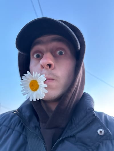 An extreme low-angle selfie captures a young person looking directly at the camera, set against a clear, bright blue sky. The individual wears a dark-colored baseball cap or beanie, a dark brown hooded sweatshirt with the hood up, and a dark blue or black puffer jacket. A prominent silver snap button and zipper are visible on the jacket. A single white daisy, featuring delicate white petals and a vibrant yellow center, is held between the person's lips or near their chin. Their eyes are wide and focused forward. Faint, thin lines, possibly utility wires, are subtly visible in the upper portion of the bright sky. The clear sky suggests a daytime setting with good weather. No specific location identifiers or text are visible in the image.