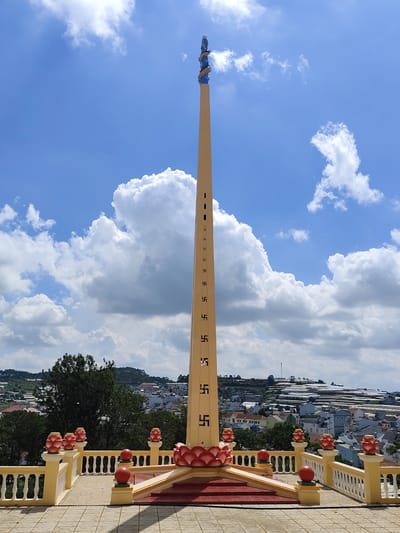 An outdoor, eye-level, medium shot captures a tall, narrow, cream-colored tower, the main subject, set against a bright blue sky filled with puffy white clouds. The tower tapers towards its peak, which is adorned with a blue and gold decorative element resembling a dragon and a small blue figure. The tower's surface is decorated with a repeating pattern of swastika symbols. In the foreground, a yellow railing with white balusters encloses a tiled platform. Decorative lotus flower sculptures and red spheres are placed along the railing. A set of red stairs with yellow trim leads up to a raised platform in front of the tower, also decorated with lotus sculptures. Behind the platform, a sprawling city unfolds with a mix of houses and buildings, many with grey or blue roofs. Green trees are scattered throughout the city, and a hillside with numerous greenhouses is visible in the distance. The scene appears to be located in the city of Lộc Quý, Vietnam, as indicated by the presence of greenhouses common in the region and the architectural style.