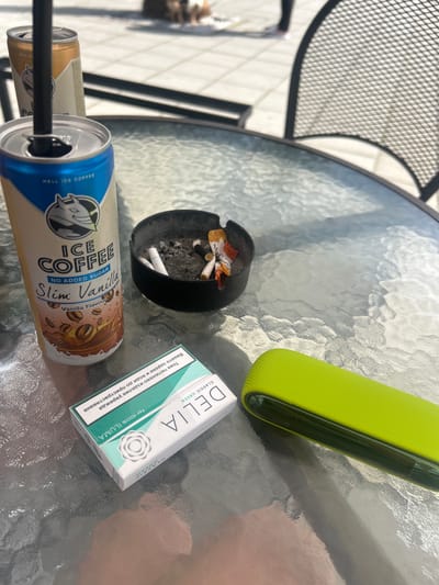 On a textured glass table top, an ashtray with cigarette butts is positioned near a pack of "Delia" Classic Green for IQOS ILUMA cigarettes. A green IQOS device rests nearby. Two cans of "Hell Ice Coffee," one blue and one gold, are visible, with a straw in the blue can. The scene appears to be outdoors, possibly in Plovdiv, Bulgaria, given the Cyrillic text on the cigarette pack. A metal chair is partially visible.