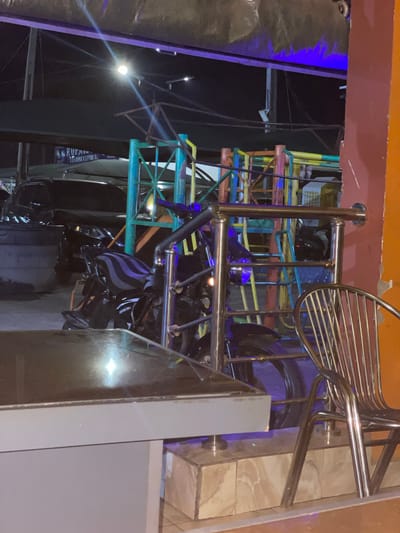 The image depicts a vibrant, multi-colored structure, possibly a piece of playground equipment, positioned within an indoor environment. In the background, a dimly lit exterior is visible, hinting at an outdoor setting. A metallic chair is situated near the foreground, with various other metal and plastic objects scattered throughout the scene. The lighting is uneven, with some areas brightly illuminated and others shrouded in shadow, creating stark contrasts. The location is Hayin Banki, Nigeria.