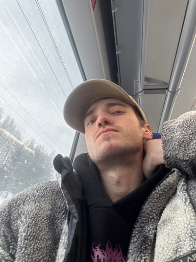 A young adult male, likely Caucasian, is depicted in a close-up selfie taken inside a public transport vehicle, presumably in the city of Redkino, Russia. He is facing the camera with a direct gaze, wearing a beige baseball cap, a black hooded sweatshirt with a partially visible purple graphic, and a mottled gray and white sherpa fleece jacket, partially unzipped. His right hand is positioned behind his neck. The interior of the vehicle features a light-colored ceiling with horizontal panels, a vertical silver support pole on the right, and a horizontal white bar with the alphanumeric inscription "474.48.SD". Through the large window occupying the left and upper portions of the frame, a winter landscape is visible, characterized by snow-covered trees and an overcast sky. Overhead wires, possibly power lines, traverse the exterior view.