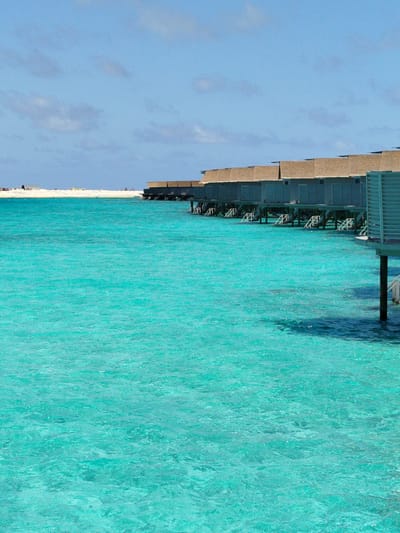 The image shows a row of overwater bungalows on the island of Gulhi Falhu, Maldives. The bungalows are constructed on stilts over turquoise water and have thatched roofs. The water is clear and shallow, with a sandy seabed visible in some areas. The sky is blue with scattered clouds. There are no people visible in the image.