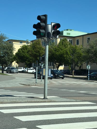 This image captures a street scene in Helsingborg, Sweden, likely during daylight hours on a clear day, given the bright sunshine and distinct shadows. The primary subjects are traffic lights and a pedestrian crossing. The traffic lights are the main focus, with a set of vertical signals visible, one of which displays a green light. Beside the pedestrian crossing, which is clearly marked with white stripes on asphalt, is a lamppost and several parked cars.
In the background, buildings with light yellow facades and green accents are visible, suggesting an urban or suburban environment. Trees with lush green foliage are scattered throughout the scene, adding a natural element to the cityscape. The perspective of the image is slightly elevated, looking down onto the street and crossing. No people are directly visible in the frame, and the overall atmosphere is calm and uneventful, consistent with a typical day in a city. The presence of the pedestrian crossing and traffic lights indicates a regulated intersection.
One vehicle's license plate is partially visible, showing "UBE 456", which is a Swedish license plate format. The overall scene depicts a mundane urban setting in Sweden, focusing on traffic infrastructure and its surroundings.