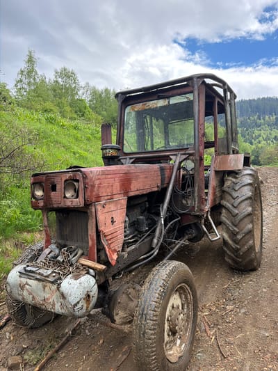 The image shows a rusty, dilapidated tractor located near Рилски манастир, Bulgaria. The tractor is missing parts and is surrounded by mud and vegetation. The environment consists of a dirt area bordering a grassy hillside with trees and a cloudy sky.