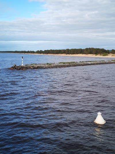 The image depicts a body of water, likely a lake or sea, with a rocky breakwater extending into it. The breakwater is lined with some vegetation and a small, white and black striped navigation aid is located at its terminus. The shoreline in the background shows a sandy beach bordered by a dense line of trees. In the foreground, a white navigation buoy floats in the water. The sky is partly cloudy. The precise location in the city of Ozernoe Ust'e, Russia, is not verifiable from the visual information alone.
