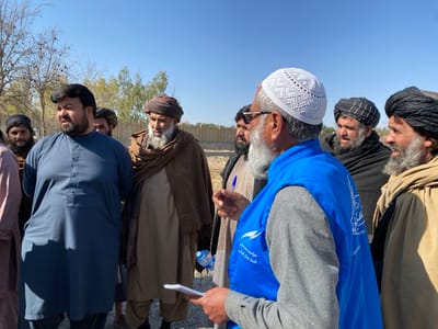 A group of men stand outdoors in an open space under a clear blue sky. Some men wear turbans while another wears a white prayer cap. A man in the foreground holds a pen and paper wearing a blue vest with lettering and a logo. The men are of varying ages with beards, dressed in traditional clothing, and appear to be listening to the man in the vest. Trees with bare branches are visible in the background.