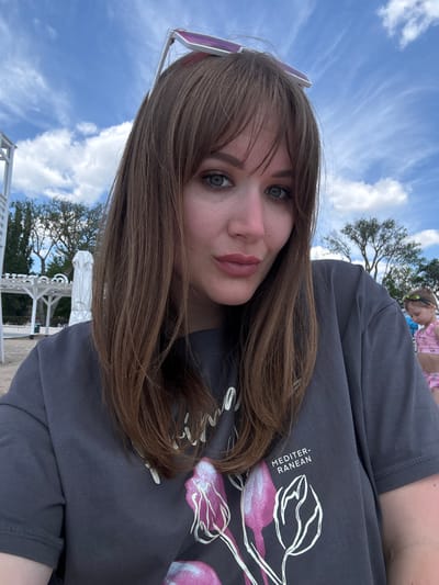 The image presents a woman with fair skin, light brown hair, and blue eyes, posing in front of a clear blue sky with scattered white clouds. She wears a dark gray t-shirt with a graphic design that includes white and pink floral elements, along with the word "MEDITER-RANEAN". The lower portion of the image reveals white structures, possibly gazebos or architectural elements, suggesting an outdoor setting near the beach. A tree is partially visible behind the structures. The location is likely Yevpatoriya, Ukraine, based on visual cues and the prompt's request. In the background, a child is wearing a pink bathing suit.