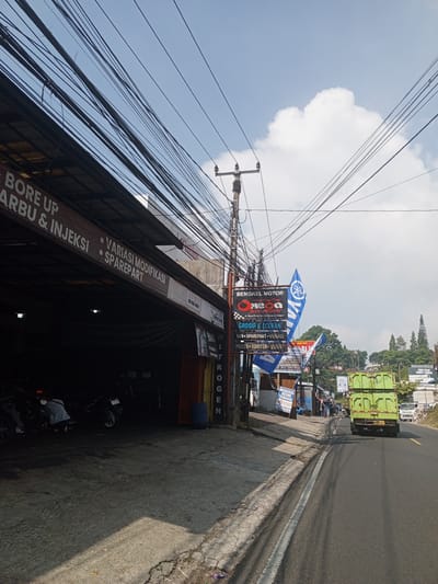 The image depicts a roadside scene in what appears to be a commercial area. In the foreground, a dark, open-fronted workshop is visible, with several motorcycles parked inside and a person working on one of them. A banner above the workshop entrance reads "BORE UP CARBU & INJEKSI VARIASI MODIFIKASI SPAREPART". Electrical wires are strung across the sky, obscuring a bright blue sky with scattered clouds.

To the right of the workshop, a road runs alongside, with a green dump truck driving away from the camera. Several signs are visible along the roadside, including one for "BENGKEL MOTOR MEGA" and another that appears to advertise motorcycle parts and services. In the background, lush green hills rise, dotted with buildings and trees. The overall impression is of a busy, functional street scene in a possibly tropical or subtropical region. The presence of "Ruwan Eliya, Sri Lanka" in the prompt does not align with any visual cues in the image, which suggests a Southeast Asian context due to the signage.