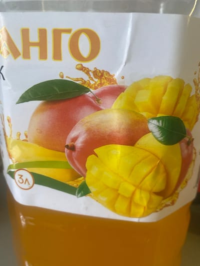 The image shows a close-up of a package of mango juice, likely a liter or more based on the "3л" marking. The packaging is predominantly white with a vibrant illustration of mangoes and mango slices. A large, stylized word, appearing to be "МАНГО" in Cyrillic, is printed in an orange and yellow gradient at the top of the package. The juice itself is a deep orange color, visible in the lower portion of the image. The image does not provide any information about the city of Mamontovo, Russia.