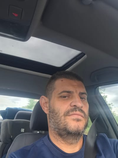 A man with short, dark hair and a beard is taking a selfie inside a car. He is wearing a dark blue sweatshirt and a seatbelt. The car has a sunroof, and the exterior appears to be overcast with some trees visible. There is a red emergency button on the ceiling of the car. The car interior includes dark gray seats with a white emblem on the headrest. The man's face is centered in the frame, with his gaze directed at the camera. The setting does not provide specific visual cues to pinpoint a location within the city of Gabarevo, Bulgaria.