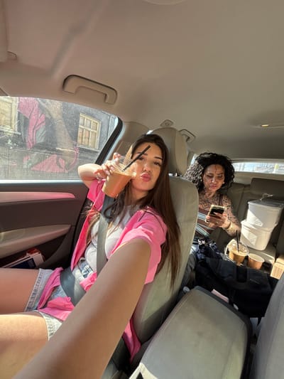 The scene is set inside a car in Rakovski, Bulgaria, illuminated by sunlight. In the front passenger seat, a young woman with long dark hair, wearing a pink open shirt over a light grey ribbed tank top and denim shorts, holds a clear plastic cup with a light brown beverage and a black straw. She is looking towards the camera with a pouting expression. A black seatbelt crosses her chest and lap.
In the rear passenger seat, a second woman with dark, curly hair and extensive tattoos visible on her arms, chest, and neck is engrossed in a gold-colored smartphone, holding it with both hands. She wears a striped top.
The car's interior features light beige headliner and seats, along with grey and lighter-toned door panels. Through the front passenger window, old buildings with faded, possibly peeling paint and multiple windows are visible, one featuring a large red and black mural or graffiti. The car window appears to have some specks or dust. On the floor behind the rear passenger seat, two white plastic storage containers are stacked, alongside a black bag and additional beverage cups (one clear plastic, one paper). A water bottle is also visible on the floor.