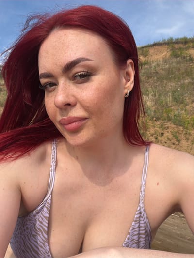 A woman with bright red hair and freckles is pictured from the chest up, outdoors. She is wearing a light-colored bikini with a zebra-like pattern. The background appears to be a sandy hill with sparse vegetation and a blue sky. The woman is looking towards the camera, with one eye slightly closed in a wink. She has on subtle makeup, including mascara and lip gloss. She is also wearing earrings: a small stud in her upper ear and a hoop in her lower earlobe. The image appears to be taken during the daytime, with natural lighting. There are no visible landmarks that would indicate a specific location within or outside the city of Ягодное, Russia.