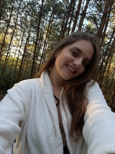 A young woman with long brown hair and a white bathrobe smiles at the camera, with a background of tall trees and a bright sky.