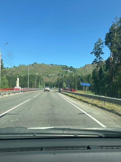 The image shows a view from a vehicle driving on a road in Souzga, Russia. The road has multiple lanes and is bordered by a guardrail and grassy areas. On the left side of the road, there is a red railing, a statue on a pedestal, and a lamppost. On the right side of the road, there are tall trees. In the distance, a mountain covered with greenery and trees is visible. A blue sign with white Cyrillic text that reads "р. КАТУНЬ" is posted on the right side of the road, indicating the Katun River. A white SUV is driving ahead on the road. The sky is clear and blue.