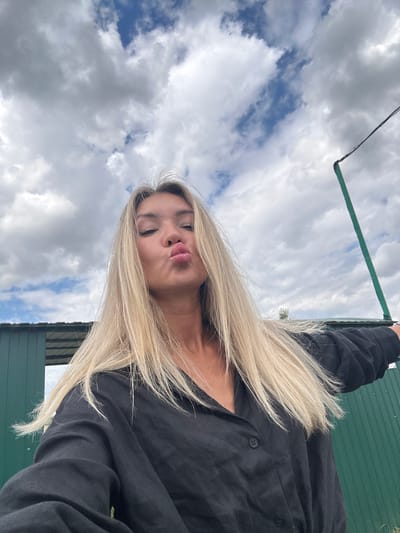 A young woman with blonde hair is captured in a selfie outdoors. She is wearing a black button-up shirt and puckering her lips. The background features a cloudy sky, a green building, and a tall green pole with a wire attached. The overall setting appears to be a rural or semi-urban area, possibly on the outskirts of Фельдмаршальский, Russia.