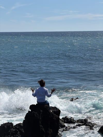 A landscape photograph shows a body of water with a rocky shoreline. A person wearing a light-colored shirt and dark pants is standing on the rocky shoreline. The person is extending their arms outwards. The waves are crashing against the rocks. The photograph was taken at Los Hervideros in Yaiza, Spain. Another person is partly visible, possibly in the water, obscured by the waves.
