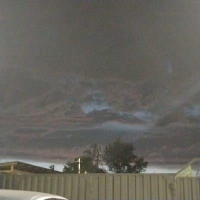 This is a medium-shot, outdoor photograph taken at night. The scene depicts a dark, overcast sky with clouds that appear to be illuminated by an unseen light source, possibly lightning or a distant city glow. In the foreground, a pale wooden fence runs horizontally across the frame. Behind the fence, there are glimpses of buildings and trees. A portion of a white car is visible in the lower left corner of the image. The overall mood is somber and atmospheric, suggesting a potentially stormy or unsettled weather condition. The location is Ekibastuz, Kazakhstan. No text is visible in the image.
